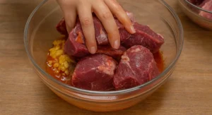 A woman’s hand placing chunks of raw beef into a glass bowl of saffron and spice marinade for Moroccan beef tagine.
