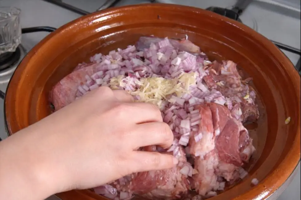 A close-up of a woman’s hand mixing raw beef with chopped onions and grated garlic inside a traditional Moroccan beef and green beans tagine on a silver gas stove.