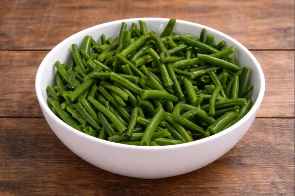 White ceramic bowl filled with cleaned and trimmed green beans on a wooden table, part of the Beef and Green Beans recipe preparation.