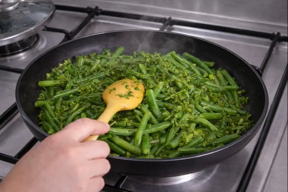 Beef and green beans. A woman’s hand stirs green beans and parsley in a black non-stick pan over a silver gas stove during the precooking phase.