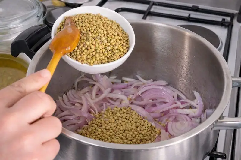 A hand pouring lentils from a white bowl into a stainless steel pot filled with sliced red onions, on a stove.