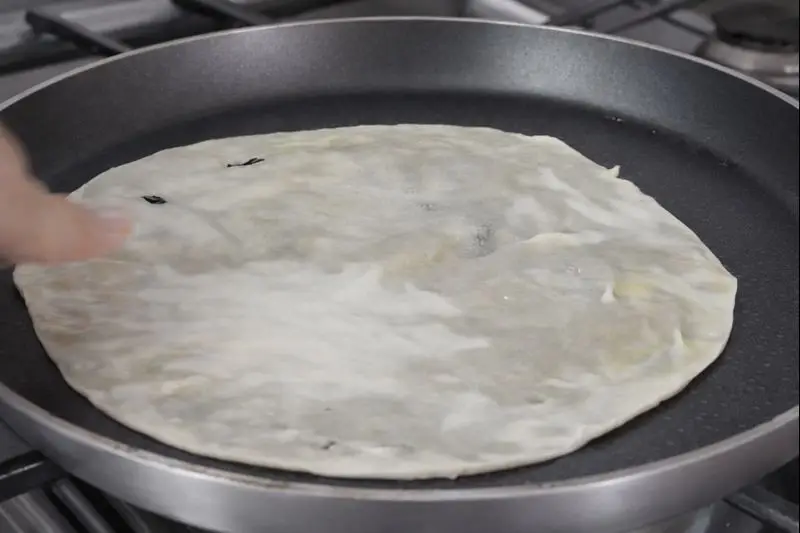 A close-up of a thin flatbread being cooked in a pan on a silver stovetop, forming traditional trid for a typical Moroccan meal called Rfissa.