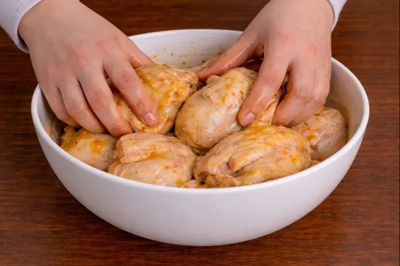 Two hands massaging marinade into coquelet chickens in a white bowl on a wooden table, preparing Rfissa, a typical Moroccan meal.