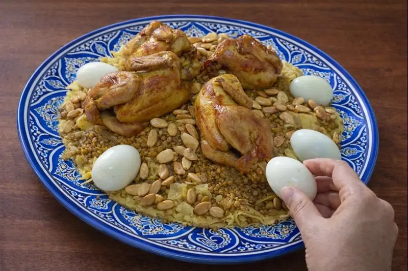 A hand picking up a hard-boiled egg from a traditional Moroccan Rfissa, a typical Moroccan meal served on a blue patterned plate.