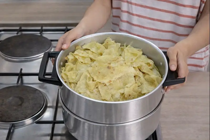 Hands holding a stainless steel couscous pot filled with torn trid flatbread pieces, positioned on a gas stove for steaming.