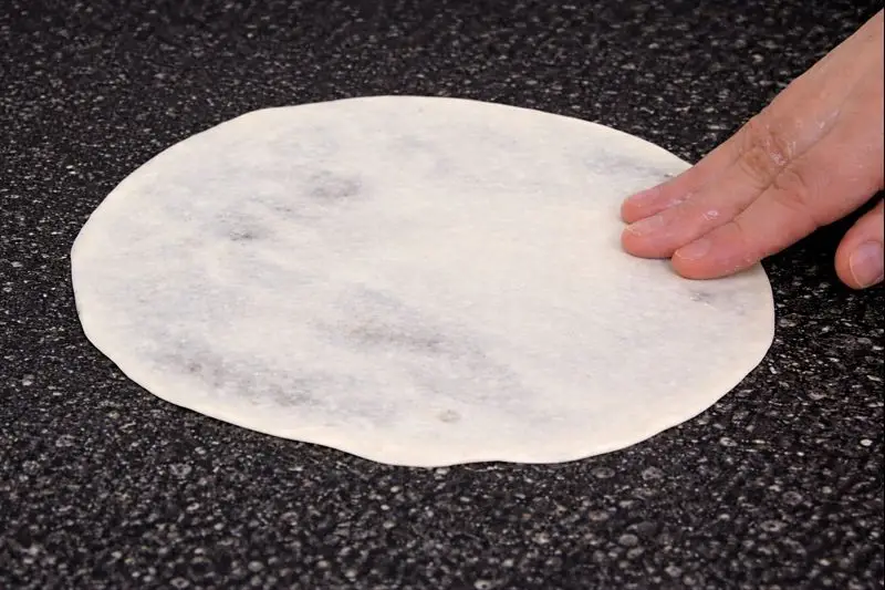 A woman's hand stretching thin dough on a black speckled countertop during the preparation of trid for Rfissa, a typical Moroccan meal.