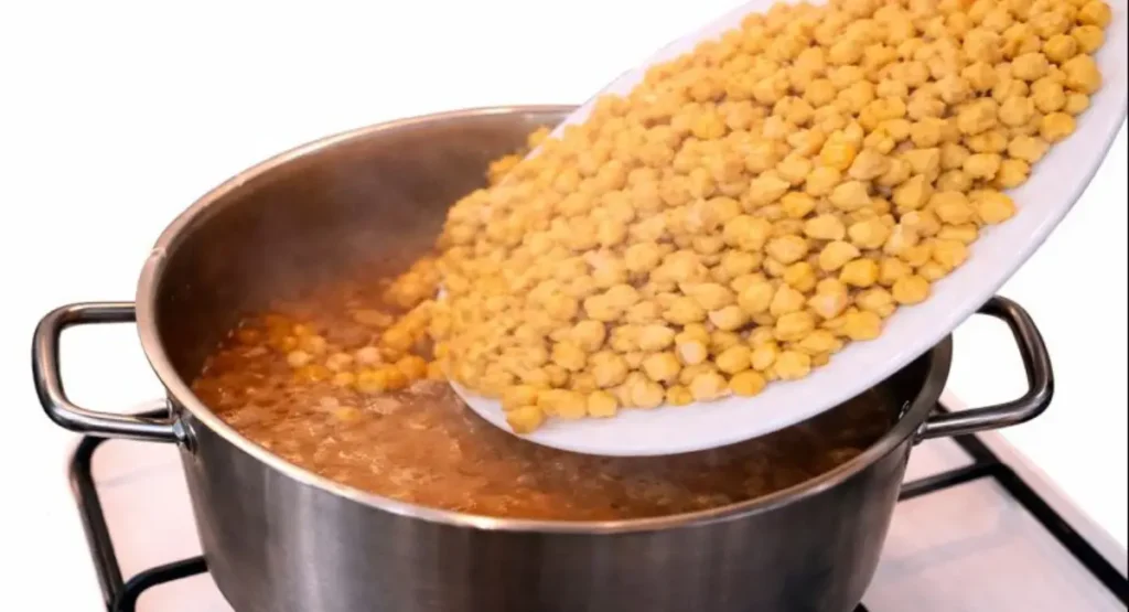 Pouring a plate of soaked chickpeas into the simmering pressure cooker filled with tender calf trotters and rich broth, marking the second cooking phase of this tagine food recipe.