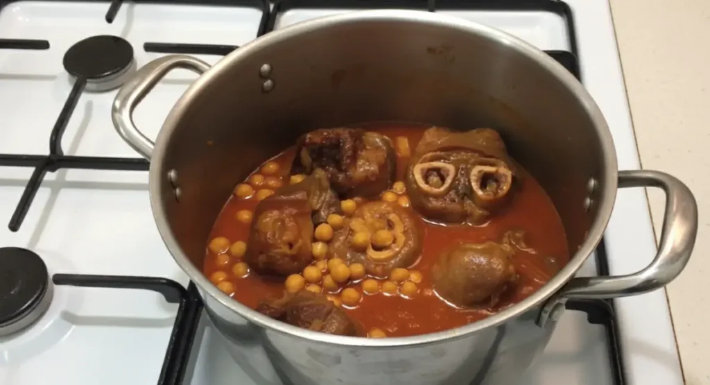 A pot of fully cooked Hargma tagine food, showing tender cow trotters and soft chickpeas swimming in a thick, reduced red sauce, ready for the final tenderness check.
