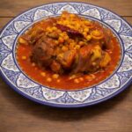 A traditional blue and white Moroccan platter filled with authentic Hargma tagine food, featuring tender cow trotters and chickpeas in a thick, sticky red sauce.