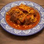 A traditional blue and white Moroccan platter filled with authentic Hargma tagine food, featuring tender cow trotters and chickpeas in a thick, sticky red sauce.