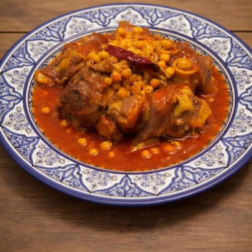 A traditional blue and white Moroccan platter filled with authentic Hargma tagine food, featuring tender cow trotters and chickpeas in a thick, sticky red sauce.