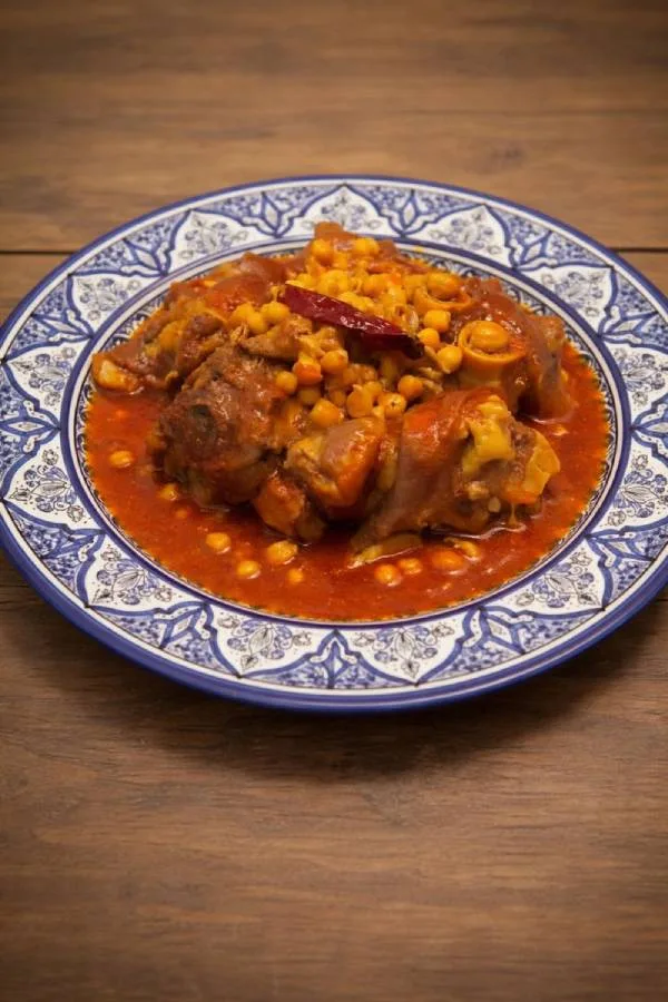 A traditional blue and white Moroccan platter filled with authentic Hargma tagine food, featuring tender cow trotters and chickpeas in a thick, sticky red sauce.
