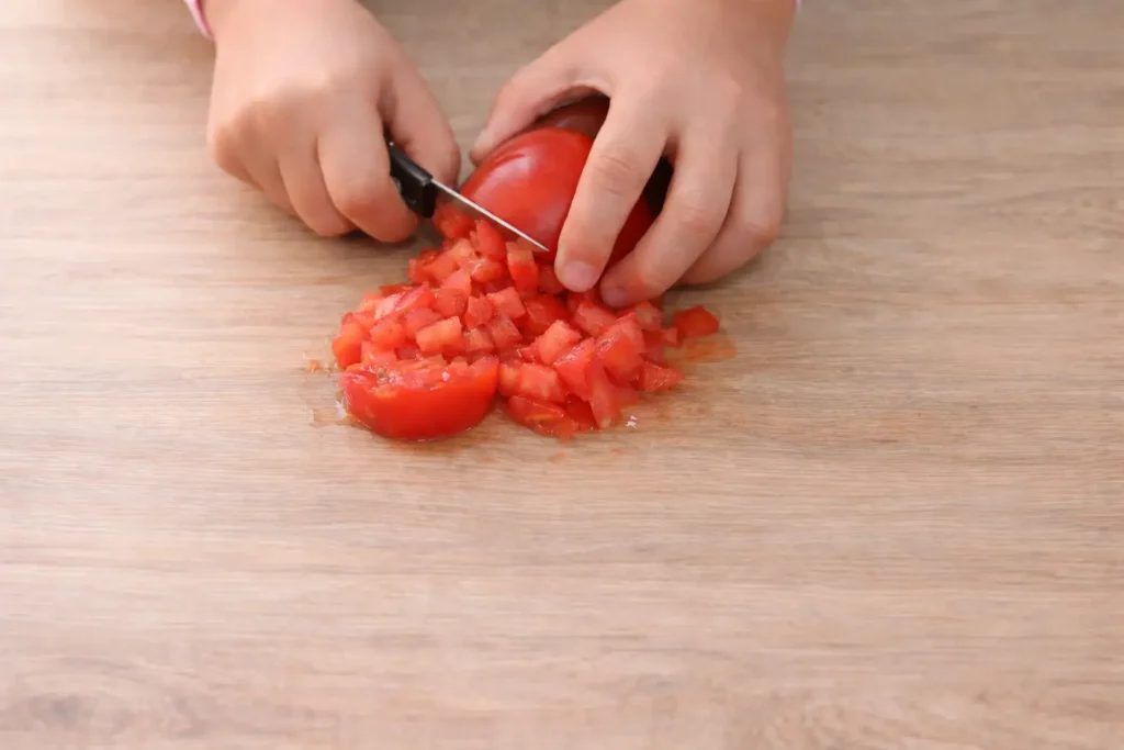 Hands cutting fresh tomatoes into cubes on a wooden table while preparing cooks illustrated zaalouk.