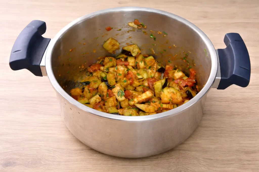 Stainless steel pot with matte blue handles cooking eggplant and tomato mixture for cooks illustrated zaalouk on a wooden table.