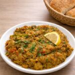 White plate of Moroccan zaalouk made with eggplant and tomatoes, garnished with fresh parsley and a lemon wedge, served on a wooden table with rustic Moroccan bread.