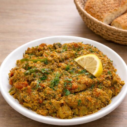 White plate of Moroccan zaalouk made with eggplant and tomatoes, garnished with fresh parsley and a lemon wedge, served on a wooden table with rustic Moroccan bread.