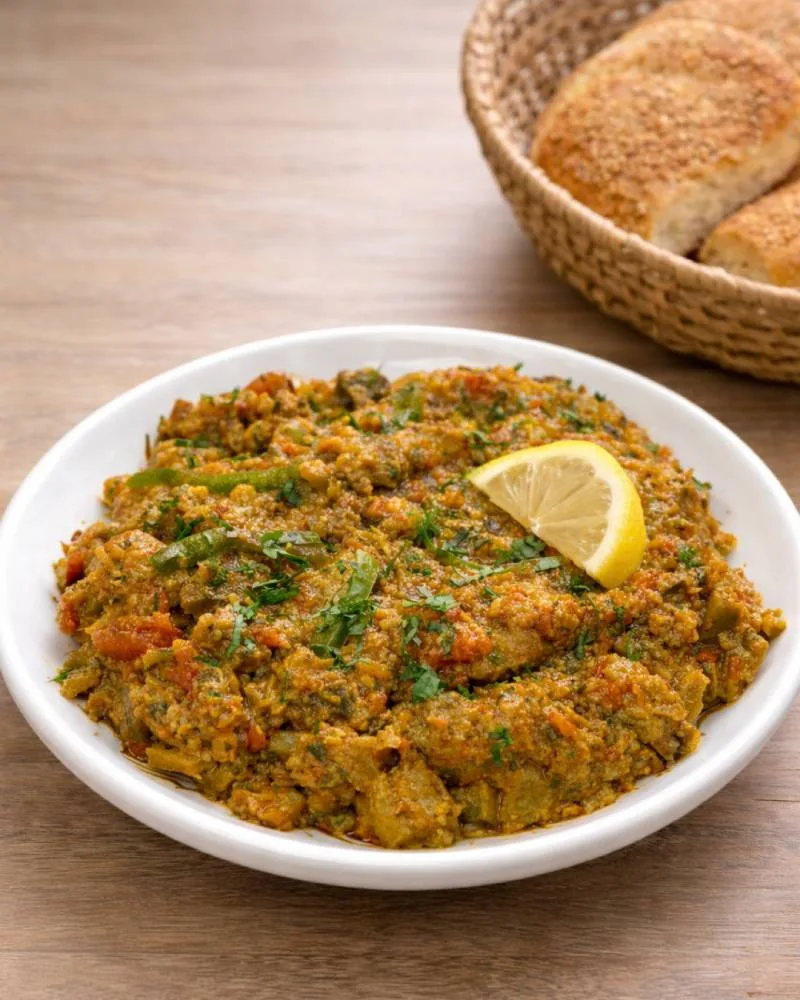 White plate of Moroccan zaalouk made with eggplant and tomatoes, garnished with fresh parsley and a lemon wedge, served on a wooden table with rustic Moroccan bread.