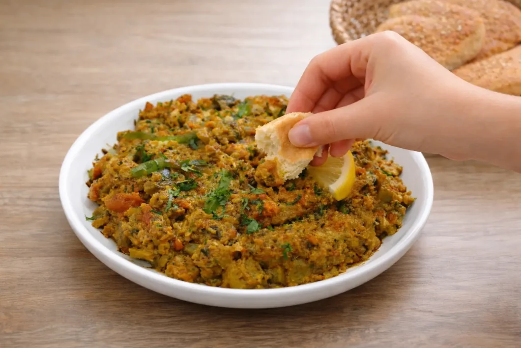 Feminine hand dipping Moroccan bread into cooks illustrated zaalouk served on a white plate with a lemon wedge.
