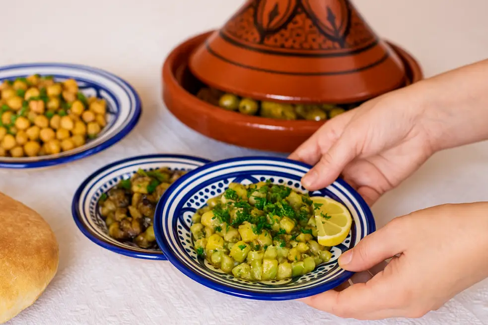 Moroccan zucchini salad. A Moroccan dining table with a central chicken tagine garnished with preserved lemon and olives, flanked by two small salad dishes, with focus on a zucchini salad being placed by a woman’s hands.