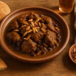 A plate of lamb Mrouzia recipe topped with toasted almonds, served with Moroccan khobz bread and a glass of tea on a wooden table