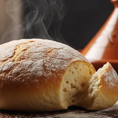 A perfectly golden-brown, freshly baked loaf of Moroccan bread, with a piece torn off to show the soft crumb.