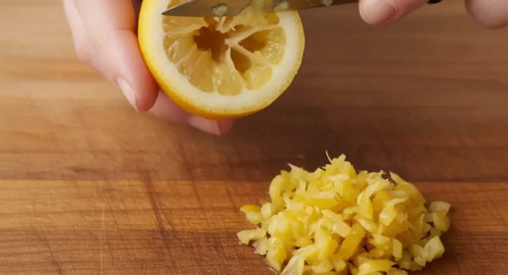 A person on a wooden cutting board demonstrating how to use preserved lemons by scraping out the pulp with a knife, with a pile of finely chopped peel ready for a recipe.